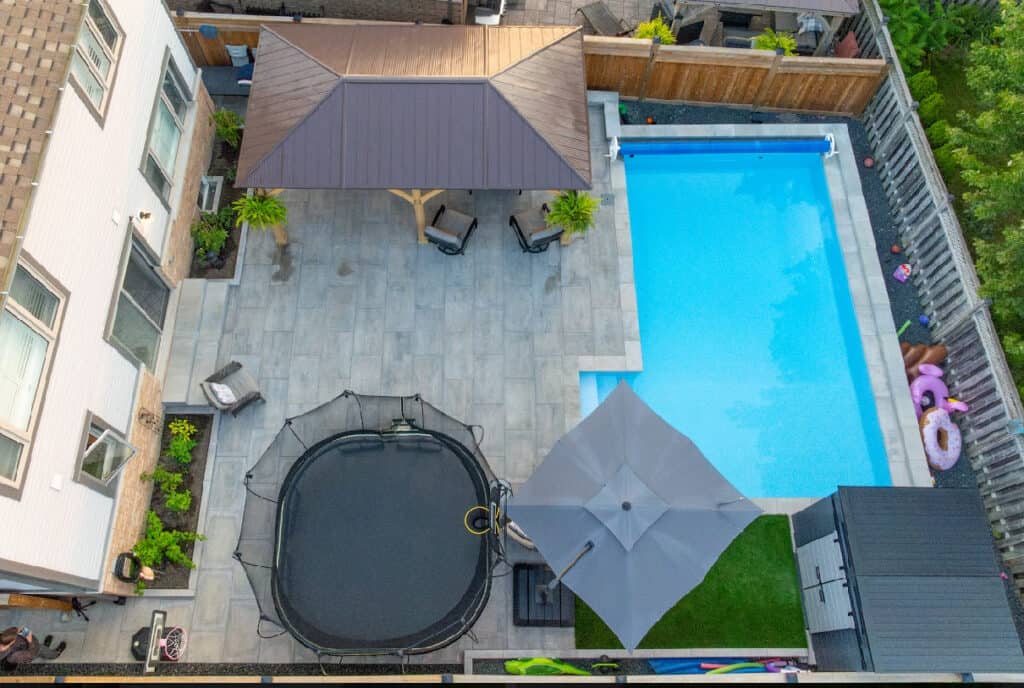 Aerial view of a landscaped backyard with swimming pool, trampoline, shaded seating area, and fencing for privacy.