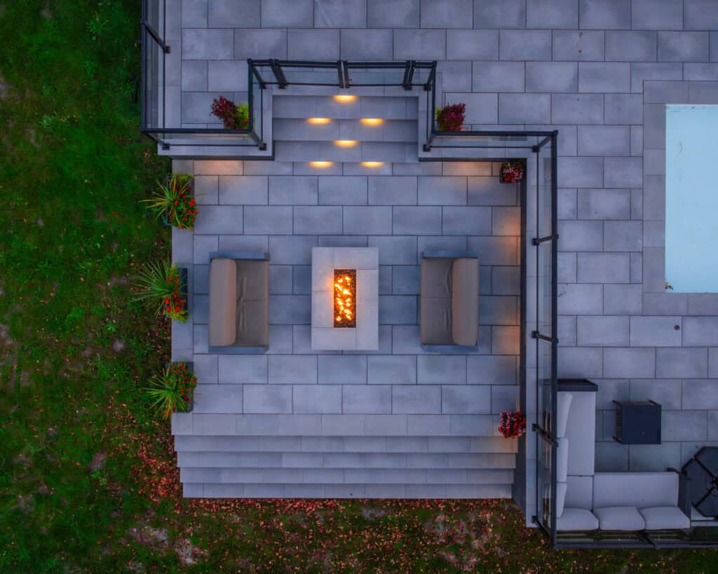 Fire pit and seating area on sleek outdoor patio with landscape lighting and greenery.